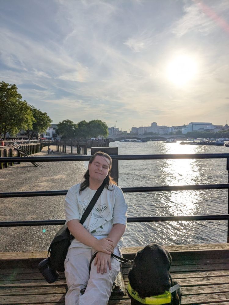 I am sitting on a wooden jetty by the Thames, with Lacey, a black guide dog wearing a yellow harness, sitting beside me. I am wearing a white t-shirt with a floral design and light-coloured trousers, and I have a black shoulder bag. In the background, there is a bridge and city buildings under a blue sky with wispy clouds. The sun is shining brightly, reflecting off the water. Trees line the riverbank on the left side of the image.