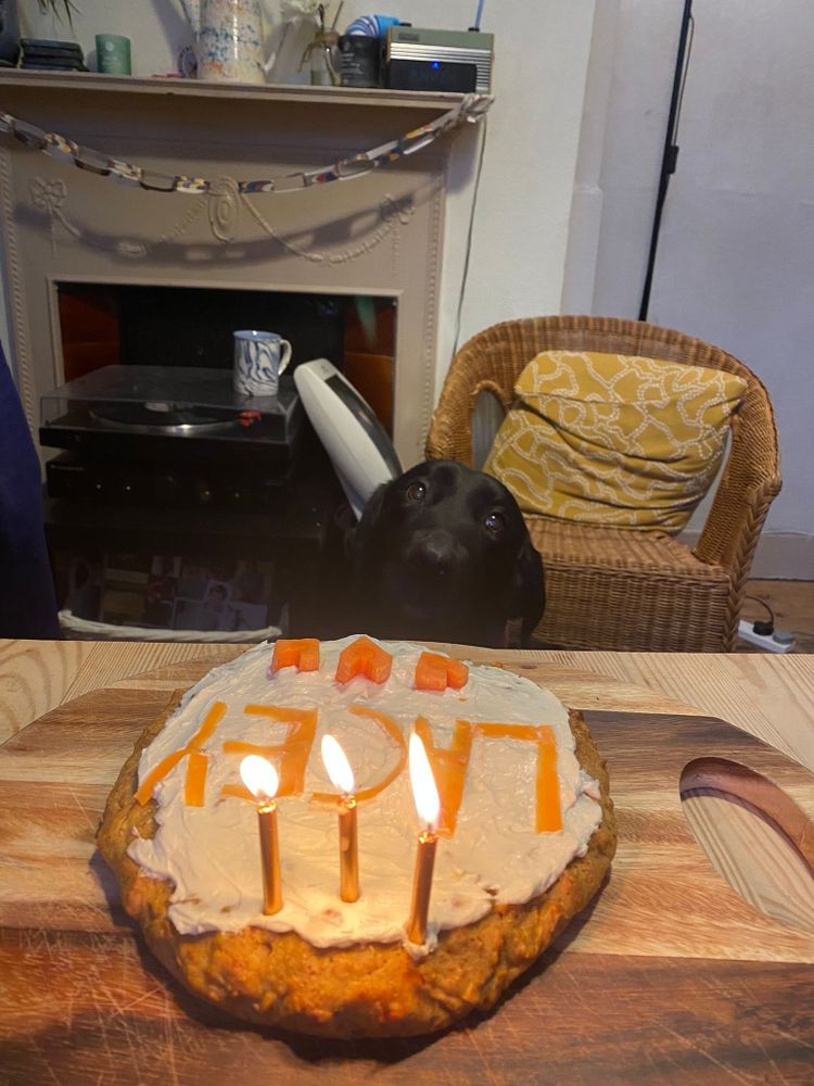 A homemade cake on a wooden cutting board with three lit candles on top. The cake is frosted with a light-colored icing and has the name "LACEY" spelled out in orange letters, made from carrots. In the background, Lacey, a black labrador retriever, has her head resting on the table, looking towards the cake.