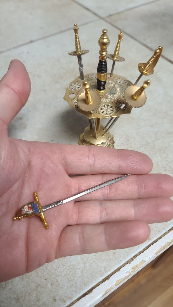 Real life photo of metal cocktail swords in a rapier style, with one in hand for size comparison. They are slightly less than the length of a finger