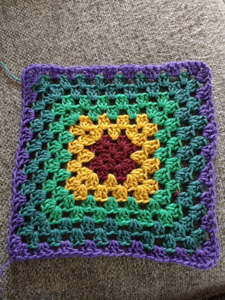 A crocheted granny square that looks like a sunflower surrounded by green with a purple border