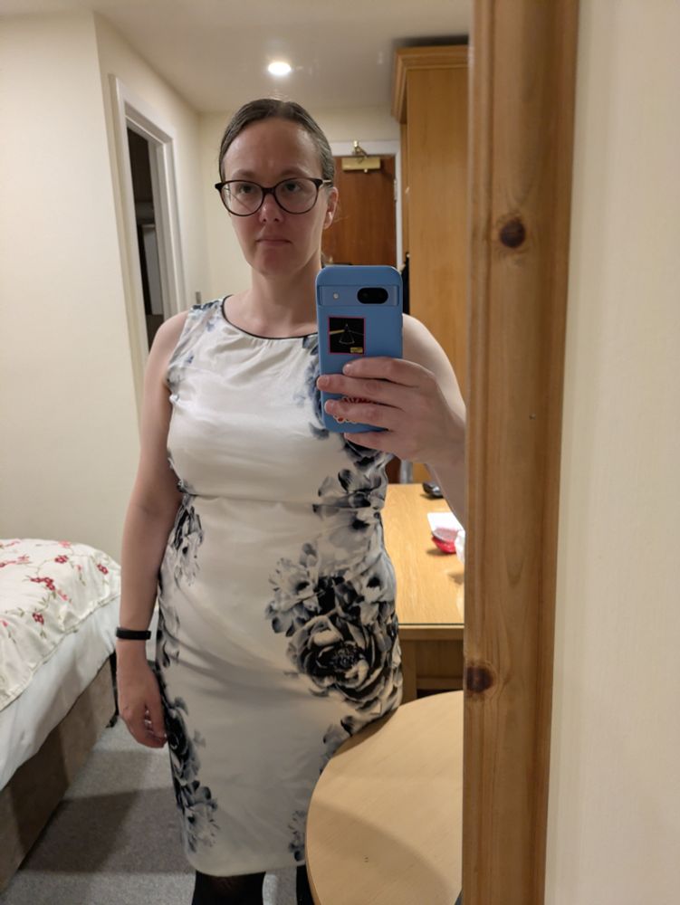 A woman with slicked backed hair and glasses wearing a white dress with black floral print down sides. 