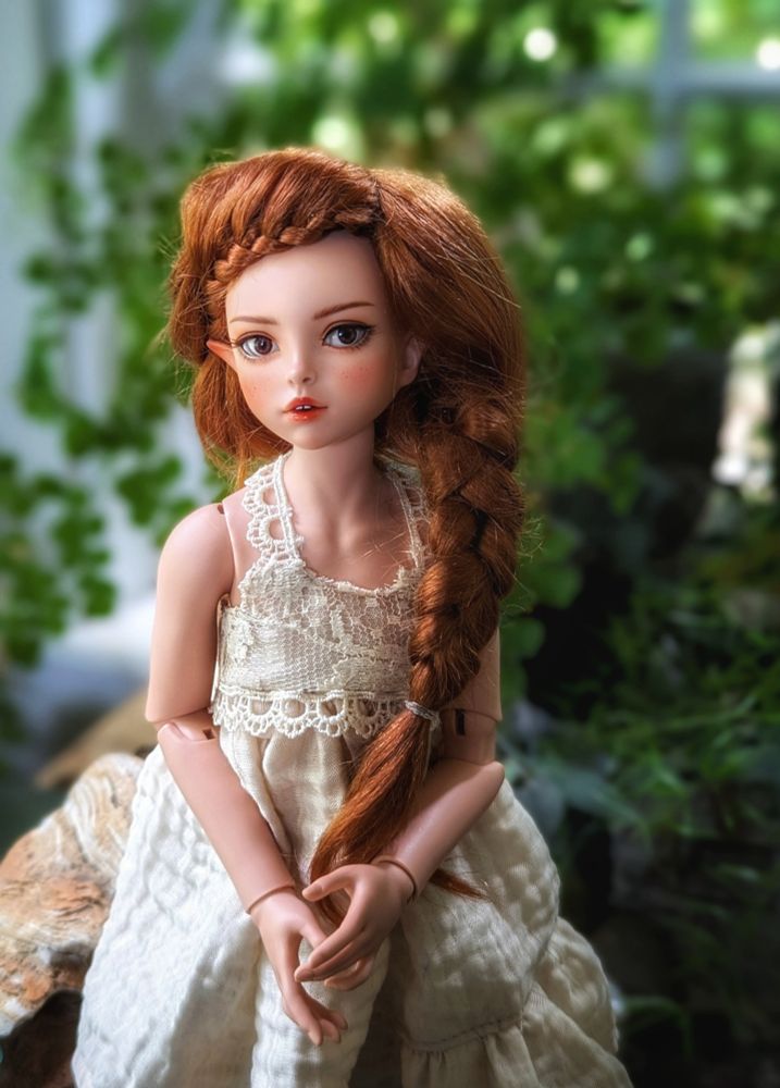 A RaccoonDoll "Angela" sculpt in rose mocha with brown eyes and braided auburn hair. She's wearing a homemade, white sundress and sitting in the indoor garden.