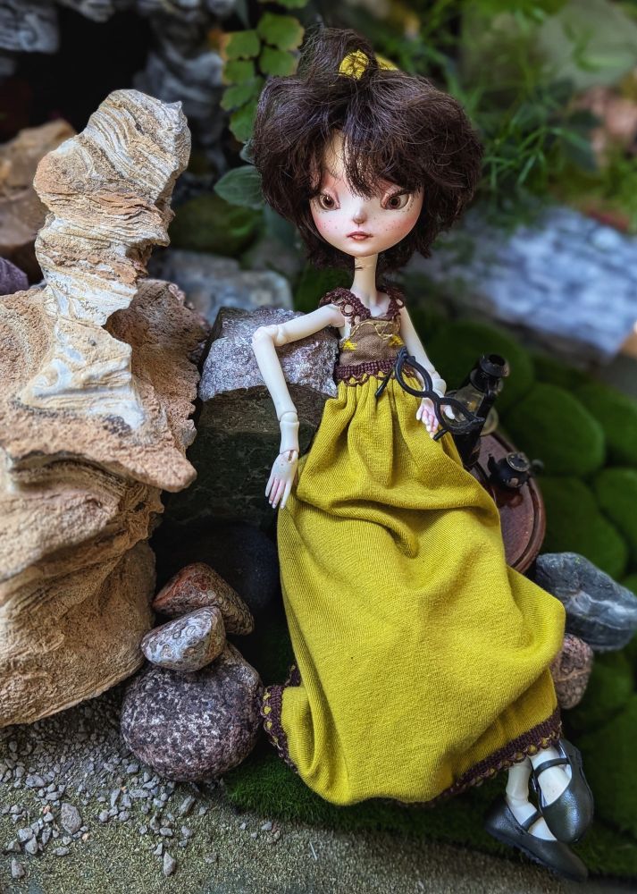 Vee, the Doll Chateau ball jointed doll, sitting in the garden, leaning on a rock, holding reading glasses in her hand.
