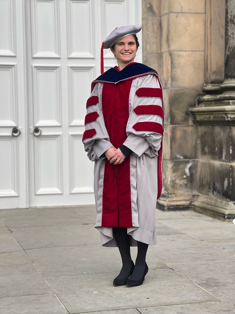 Andrea Burke stands outside a University building at St Andrews in some epic grey and red academic robes with a fun wee hat on top! 