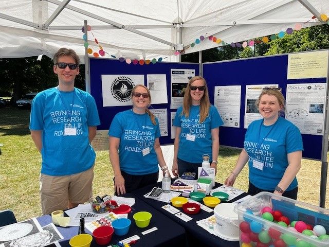 Four people in "Bringing research to life" T-shirts standing at a Southampton Clinical Trials Unit information stand, with lots of colourful science activities, posters and leaflets.