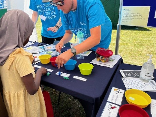 A young girl in a head scarf playing an early diagnosis research activity involving small plastic people, coloured bowls and a UV torch, with a researcher from the Southampton Clinical Trials Unit.