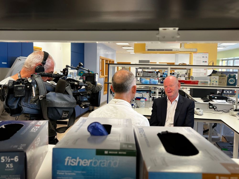 A man in a suit being interviewed by a journalist and TV camera operator in a laboratory