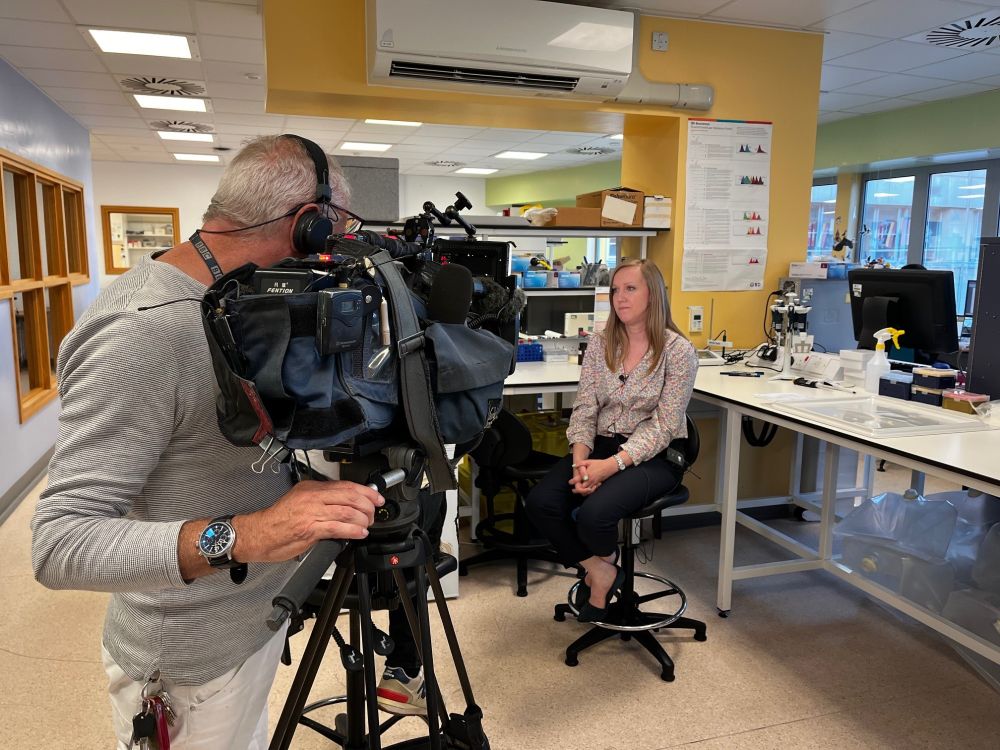 A woman in front of a TV camera operator being interviewed in a laboratory