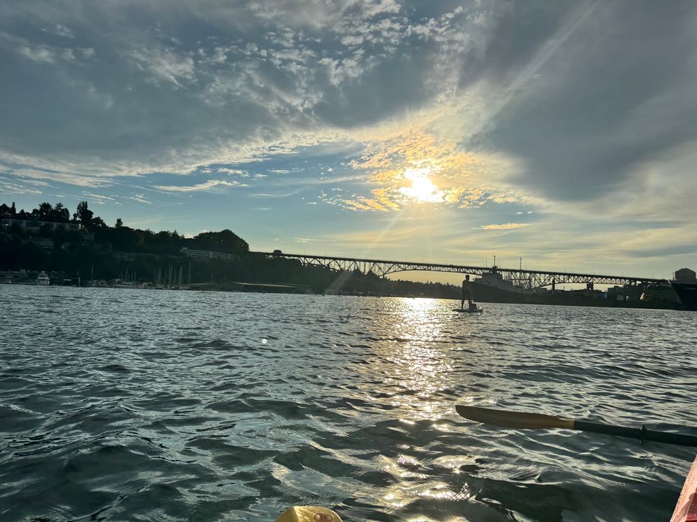 Bobbing in a kayak on a lake looking west towards a setting sun during “golden hour”. A bridge goes across the lake and a paddle boarder can be seen. The sky is golden and blue with some grey clouds