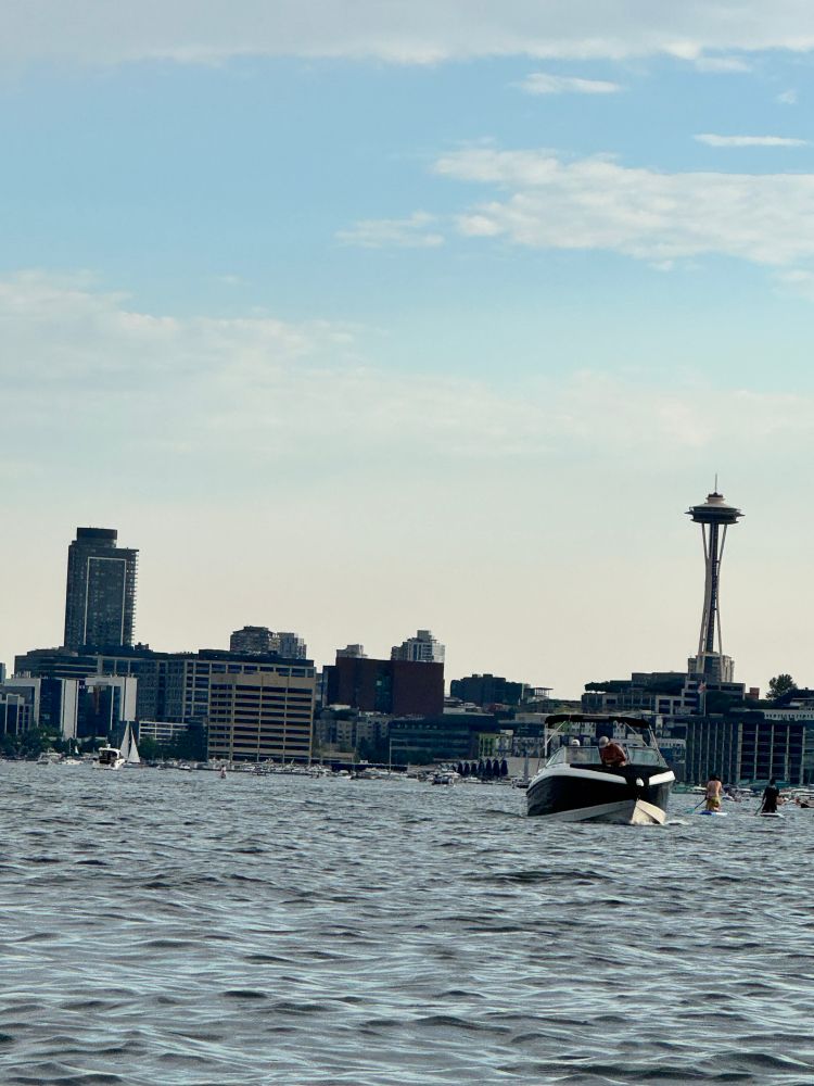 On a kayak on the lake and the Seattle skyline with the Space Needle and other buildings. There are boats and paddle boarders on the lake. The sky is blue with low clouds 