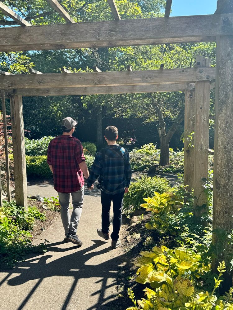 Two men, one wearing a baseball cap and a red flannel, the other in a blue flannel, holding hands as they walk down a path in a garden after an informal but sweet wedding ceremony 