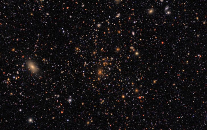 A glittering tapestry of thousands of galaxies in varying sizes, shapes, and colors. At center is a distinct collection of galaxies, all of a similar orange color.