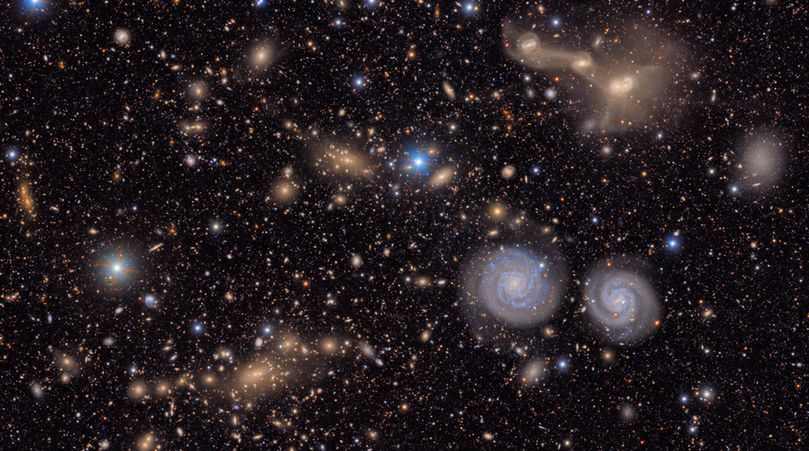 A sprawling, textured field of galaxies scattered across the deep black of space. It is filled with the delicate smudges and glowing cores of galaxies of many shapes, sizes and colors, as well as the bright multi-colored points of stars. To the lower left is a region filled with the hundreds of golden glittering gems of a distant galaxy cluster. In the foreground, below and right of center, two blue spiral galaxies look like eyes beneath the entangled mass of a triple galaxy merger in the upper right. A few bright blue points of foreground stars pierce the glittering tapestry.

All throughout the image, thousands of galaxies gather in clusters or are spread throughout, like glittering gems strewn on a table. Some are sharp-edged and spiral, like coiled ribbons; others round and diffuse, like polished pebbles. Still others are just smudges of various colors against the black of space. The background is peppered with pinpoint stars in reds, yellows, and blues, crisp against the velvet black.