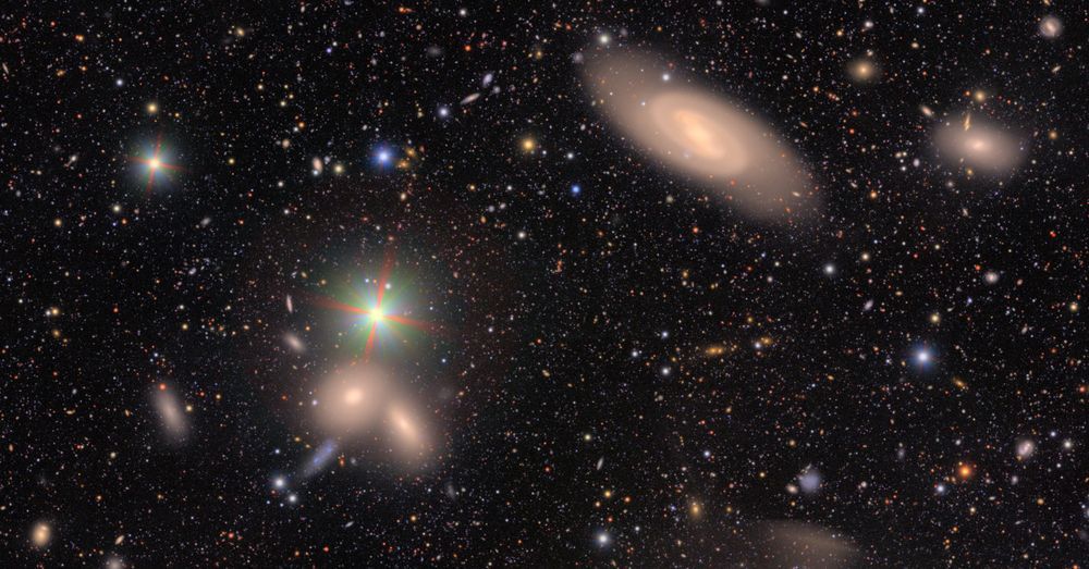 A field of galaxies of many shapes, sizes and colors, as well as the bright multi-colored points of stars. At center is a spiral galaxy peppered with tight, knotted clumps of vivid blue, young stars. In the background, hundreds of galaxies of many shapes are just smudges of various colors against the black of space.
