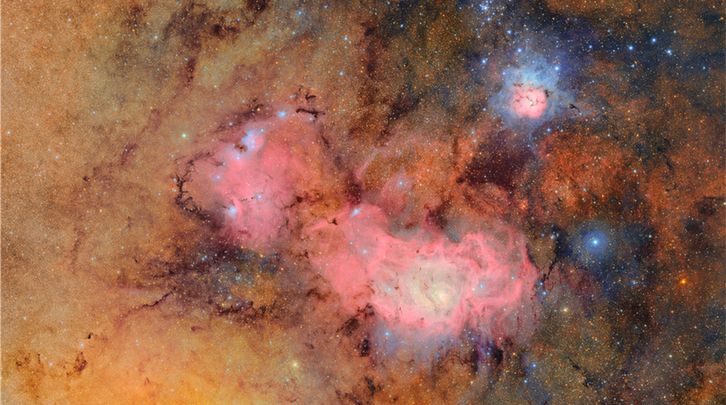 A cosmic tapestry of glowing tan and pink gas clouds with dark dust lanes. In the upper right, the Trifid Nebula resembles a small flower in space. Its soft, pinkish gas petals are surrounded by blue gas, and streaked with dark, finger-like veins of dust that divide it into three parts. It radiates a gentle, misty glow, diffuse and soft like the warmth of breath on a cold hand. To the lower left, the much larger Lagoon Nebula stretches wide like a churning sea of magenta gas, with bright blue, knotted clumps sprinkled throughout where new stars are born. Both nebulae are embedded in a soft tan backdrop of gas that is brighter on the left than on the right, etched with dark tendrils of dust and sprinkled with the pinpricks of millions of stars.
