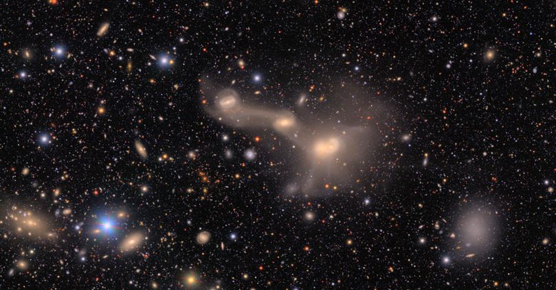 A complex entanglement of three interacting galaxies, bright cores lined up left to right and connectied by spiderweb-like tendrils. The background is black, sprinkled with countless galaxies and stars of varying colors and sizes