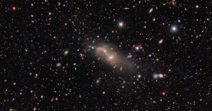 Two interacting galaxies at center, bright cores lined up upper left to lower right and connected by spiderweb-like tendrils. The background is black, sprinkled with countless galaxies and stars of varying colors and sizes.