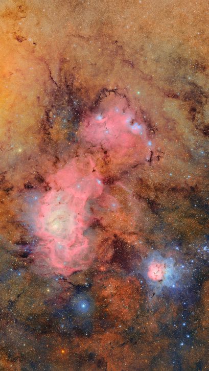 A tapestry of glowing tan and pink gas clouds with dark dust lanes. In the lower right, the Trifid Nebula resembles a small flower in space. Its pink gas petals are surrounded by blue gas, and streaked with dark veins of dust. To the upper left, the larger Lagoon Nebula stretches wide like a churning sea of magenta gas, with bright blue, knotted clumps sprinkled throughout
