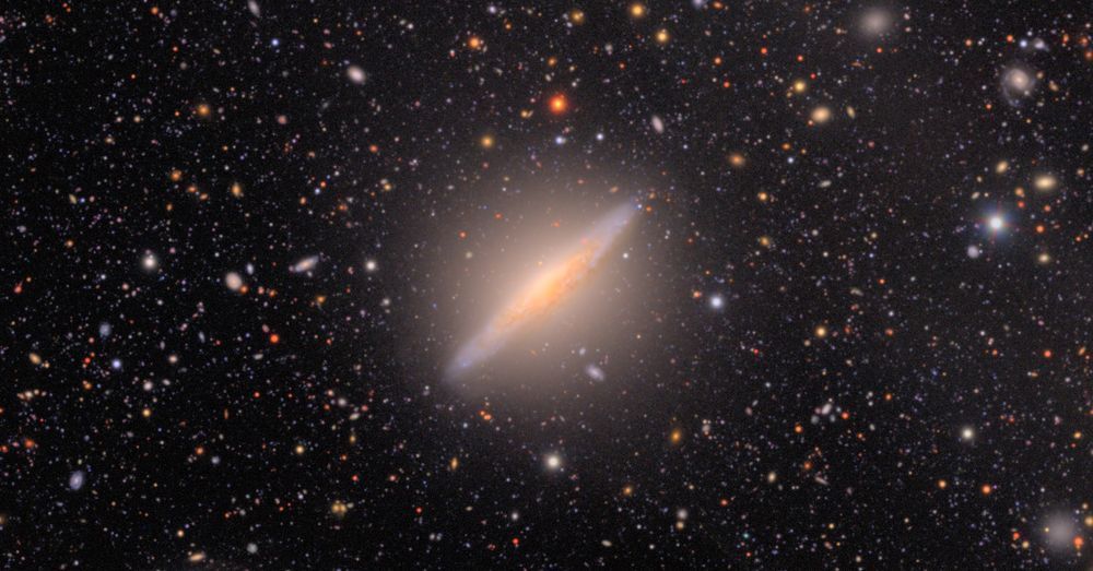 Edge-on spiral galaxy angled pointing lower left to upper right. It's glowing with a bright core that's cut with the dark veins of dust clouds. The background is peppered with a myriad of colorful stars and distant galaxies in black space.