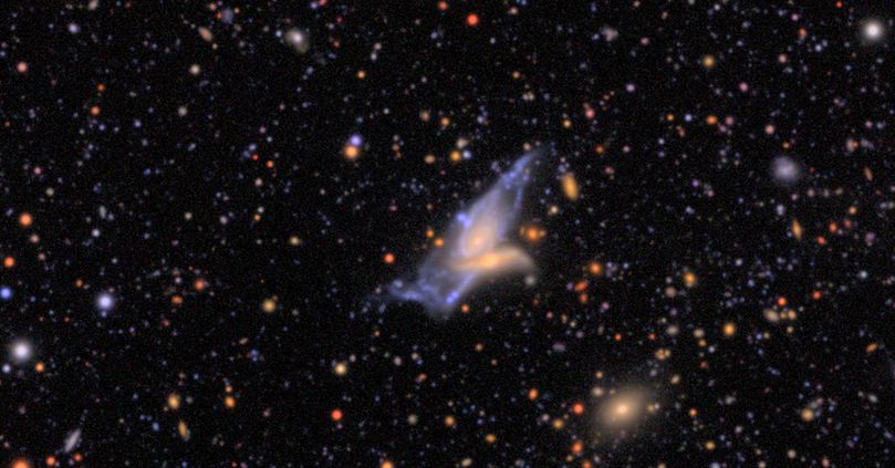 Two interacting galaxies, one larger and blue and the other smaller and more yellow. The background is sprinkled with stars and galaxies of varying colors and sizes.