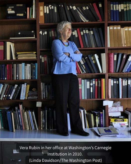 An elderly Vera Rubin with short white hair and blue shirt stands on top of a desk in front of a wide shelf full of books.
