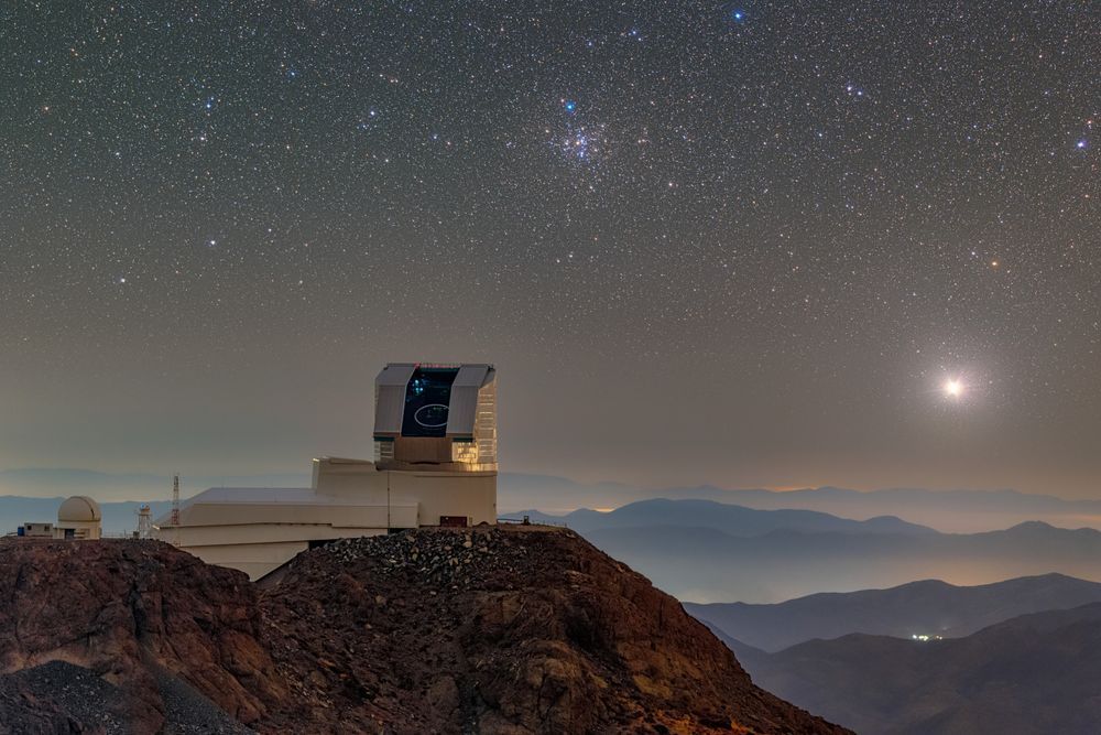 Rubin Observatory perched atop its mountain summit site on Cerro Pachón. The observatory is boot-shaped at lower left with long white service building extending left and silver dome sticking up. The dome's vertical slit is open, revealing the telescope in the blackness within. The night sky is peppered with the pinpricks of stars, and the Moon shines like a spotlight at right. The Little Beehive Cluster of stars is a denser collection of pinpricks to the upper right of the observatory.