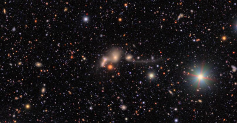 A complex yet compact collection of four interacting galaxies. The three galaxies to the left are virtually indistinguishable, with only their cores somewhat standing out. In the "elbow" of these three galaxies is a larger red star. Finally, the fourth galaxy is connected to the three by a barely visible tendril, while a second long tendril extends to the right. The background is black, sprinkled with countless galaxies and stars of varying colors and sizes.