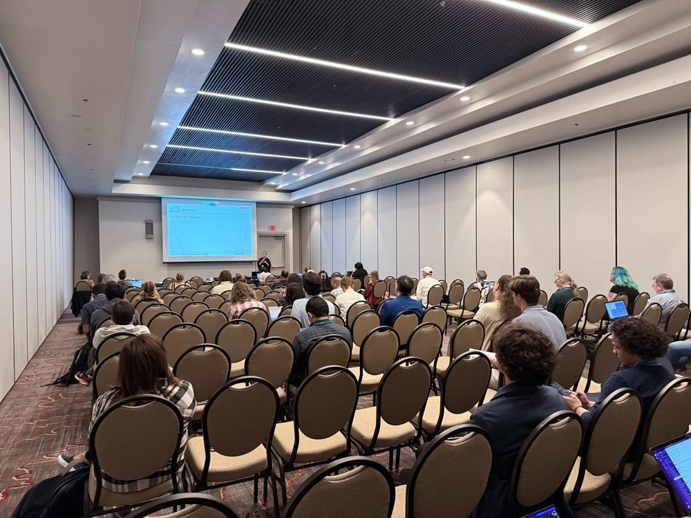 A view from the back of a conference room, with presentation slides on screen at the front of the room, and people occupying rows of seats.