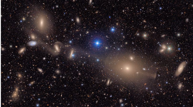 A sprawling, textured field of galaxies scattered across the deep black of space. It is filled with the delicate smudges and glowing cores of galaxies of many shapes, sizes and colors, as well as the bright multi-colored points of stars. The image focuses on a collection of interacting galaxies connected by delicate streams of stars. At top center lies a large elliptical galaxy that is dense and smooth, like a polished stone glowing with golden light. Like delicate spider silk or stretched taffy, these stellar bridges link the large elliptical to the few larger galaxies beneath, evidence of past collisions.

All throughout the image, thousands of galaxies gather in clusters or are spread throughout, like glittering gems strewn on a table. Some are sharp-edged and spiral, like coiled ribbons; others round and diffuse, like polished pebbles. Still others are just smudges of various colors against the black of space. The background is peppered with pinpoint stars in reds, yellows, and blues, crisp against the velvet black. 