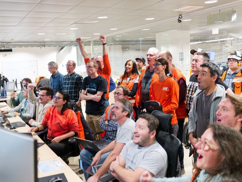 Around two dozen people sit and stand at a long desk with computer monitors out of view. Several people are smiling or have their mouths open in amazement. One person in the back has their arms raised in celebration.