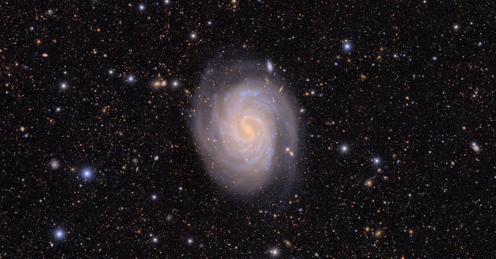 A face-on spiral with a bright core and sweeping arms knotted with blue clusters of star-forming regions. The background is peppered with a myriad of colorful stars and distant galaxies in black space.