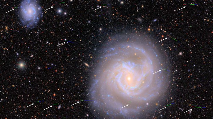 A large spiral galaxy with golden core and arms knotted with the blue clumps of stars forming sits against a background of velvet black peppered with galaxies of various shapes, sizes, and colors. A few dozen multi-colored streaks photobomb the spiral galaxy, with white arrows pointing at them. The streaks are like sprinkles across the view, just a tiny fraction of the width of the large galaxy. From left to right, the streaks are red, then green, then blue.