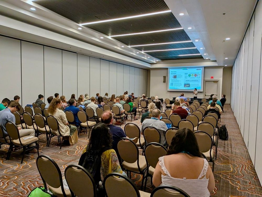 A view from the back of a conference room, with presentation slides on screen at the front of the room, and people occupying rows of seats.