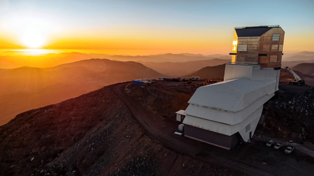 Rubin Observatory in a golden sunset, with its silver dome glinting in the light of the setting Sun. We see the observatory's long white service building and vertical silver dome such that the white service building appears pointed slightly to our left. To the left, the brilliantly setting Sun glows bright orange, illuminating the landscape in a golden glow. The Chilean desert mountain ridges become more and more opaque as they recede into the distance, creating a layered look.