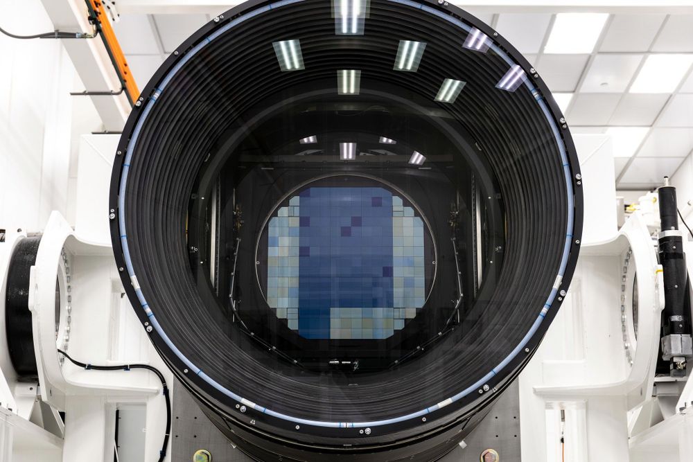 A view looking into the LSST Camera's huge opening. The camera opening is a large black donut shape, with reflections of ceiling light revealing the presence of glass lenses. In the center of the opening is the camera's focal plane detector, which is made of 189 square CCD chips arranged in a roughly square shape. The camera is suspended on a white metal frame with white handrails. The completely white room gives the image an overall sterile feel.