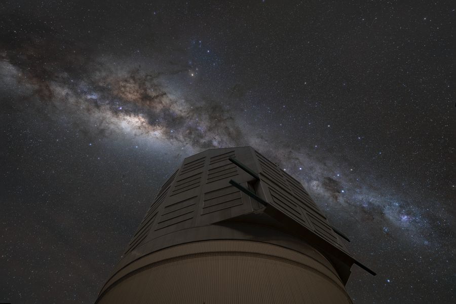 The glowing band of the Milky Way cuts across the night sky above a silver observatory dome from upper right to lower left. Dark tendrils of dust weave across the dense areas of stars of the Milky Way. The pinpricks of stars dot the rest of the sky.