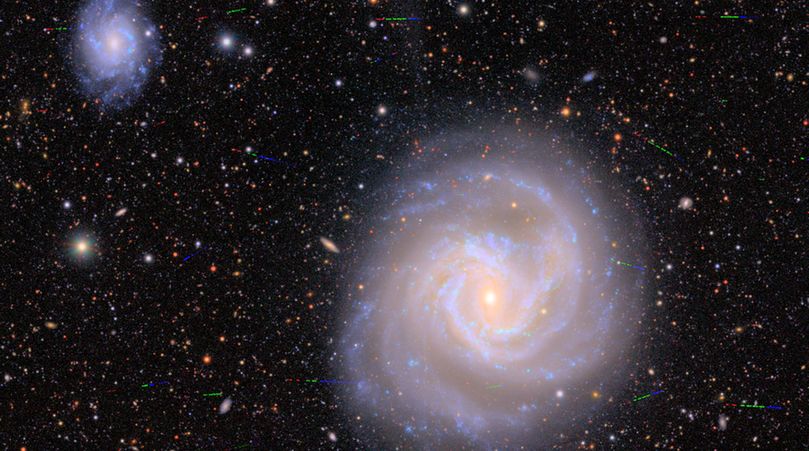 A large spiral galaxy with golden core and arms knotted with the blue clumps of stars forming sits against a background of velvet black peppered with galaxies of various shapes, sizes, and colors. A few dozen multi-colored streaks photobomb the spiral galaxy. The streaks are like sprinkles across the view, just a tiny fraction of the width of the large galaxy. From left to right, the streaks are red, then green, then blue.
