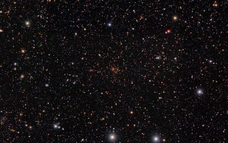 A glittering tapestry of thousands of galaxies in varying sizes, shapes, and colors. At center is a distinct collection of galaxies, all of a similar red color.