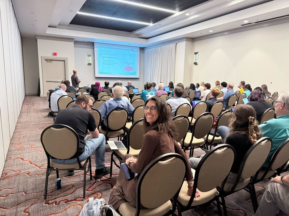 A view from the back of a conference room, with presentation slides on screen at the front of the room, and people occupying rows of seats. The person closest to the camera is turned around and smiling.