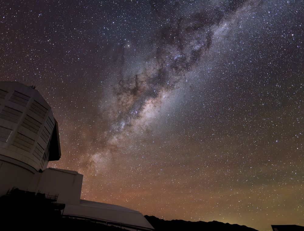 The glowing band of the Milky Way emanates from Rubin Observatory's boot-shaped silhouette at lower left. The brighter, dense stars of the Milky Way are crisscrossed by clouds and lanes of dark dust. The lower left horizon has an orangey hue, while the pinpricks of stars speckle the rest of the sky.