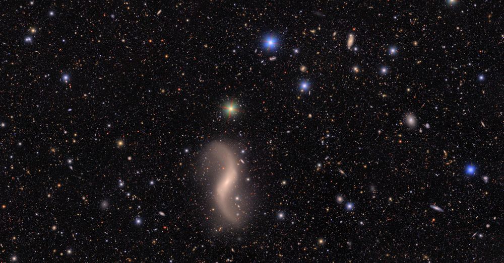 A field of galaxies of many shapes, sizes and colors, as well as the bright multi-colored points of stars. At top right is a large galaxy with two whirling spiral arms wrapping around like a hug. At bottom left, two smooth elliptical galaxies appear near each other, like champagne glasses in the midst of a cheers. A bright star above the pair has red, green, and blue diffraction spikes emanating out. In the background, hundreds of galaxies of many shapes are just smudges of various colors. 