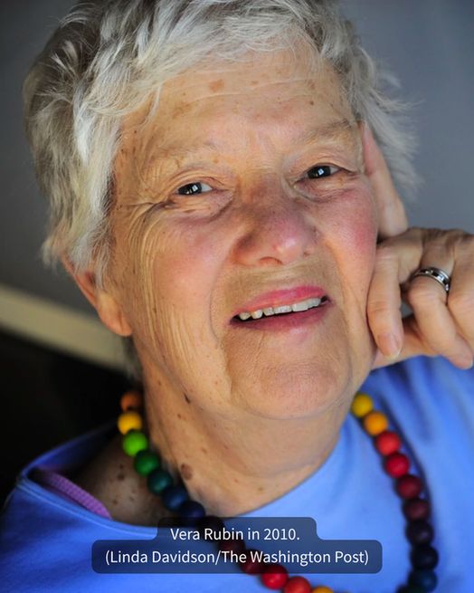 An elderly Vera Rubin with short white hair and blue shirt smiles at the camera.