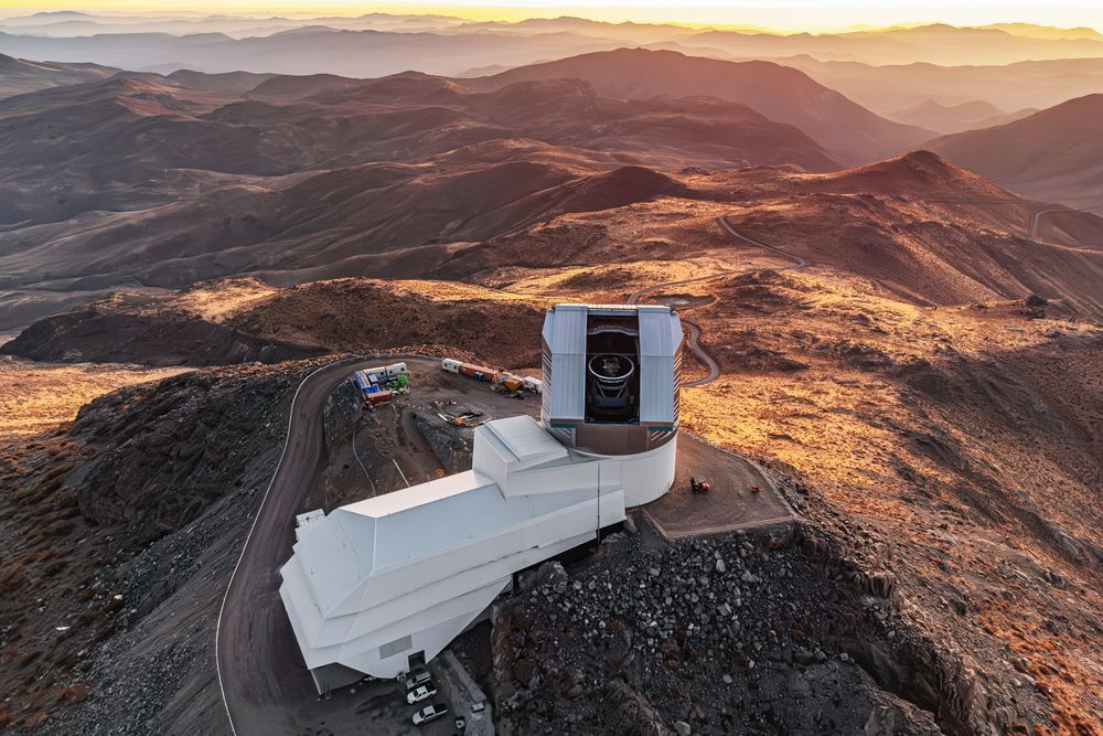 Aerial view of a boot-shaped observatory with long white service building and an open dome revealing its telescope atop a rocky mountain during golden hour with distant hills. Credit: RubinObs/NOIRLab/SLAC/NSF/DOE/AURA/T. Matsopoulos