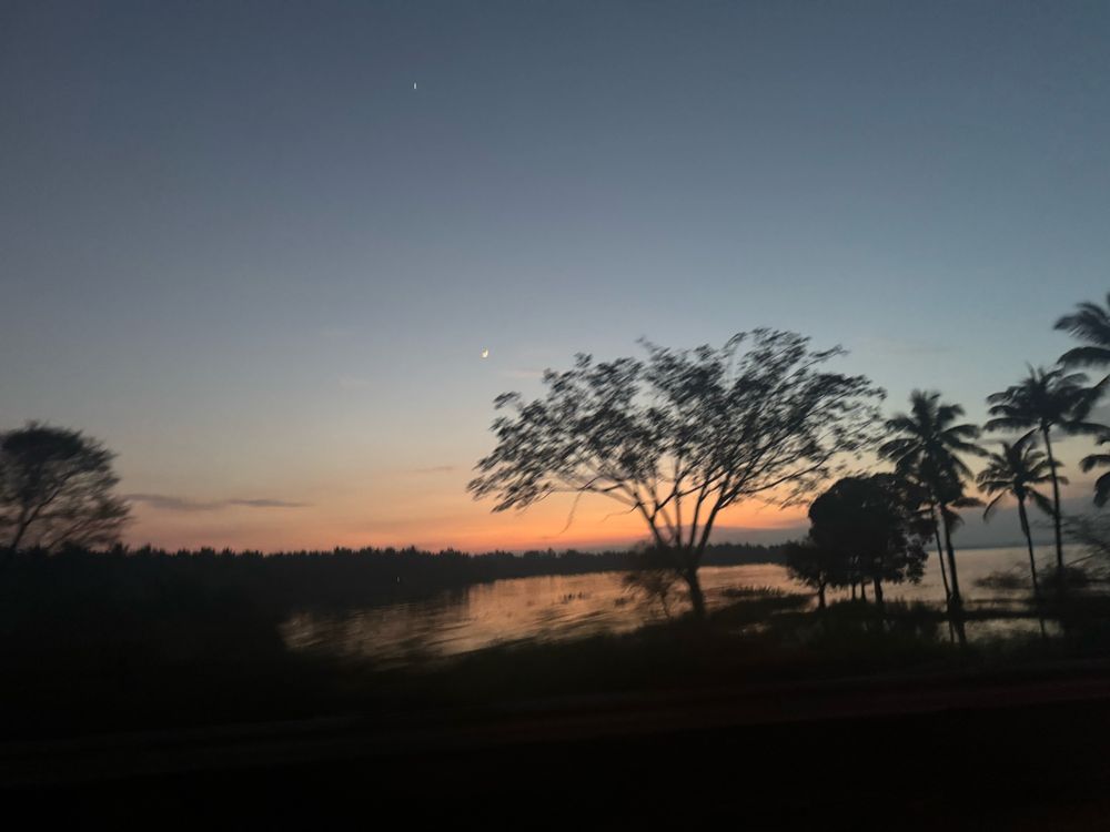 The image captures the serene backwaters of the Krishna Raja Sagar (KRS) Reservoir during sunset. Silhouettes of trees, including palms and other foliage, stand gracefully against the water, reflecting the fading hues of the evening sky. The gradient sky transitions from soft orange and pink near the horizon to a deeper blue higher up, with a crescent moon and a single bright star visible. The tranquil water and darkened landscape create a peaceful and atmospheric scene, highlighting the beauty of the reservoir at dusk.
