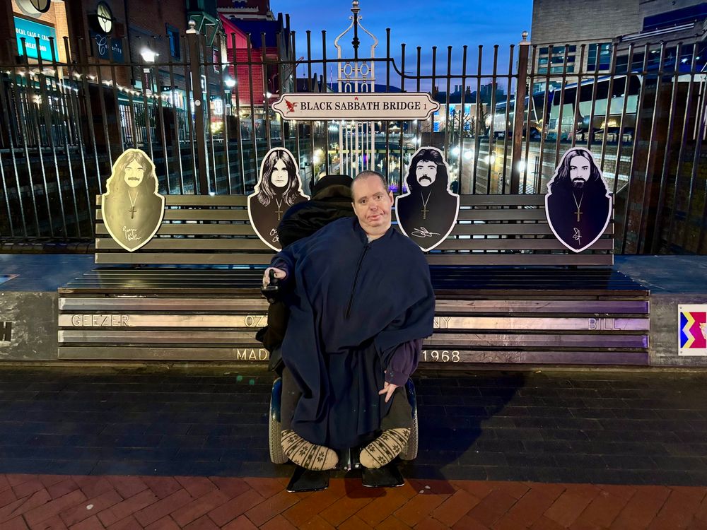 Photo of a man in a motorised wheelchair sitting in front of a bench on a bridge. The bench features photographs of the four original members of the British rock group Black Sabbath.