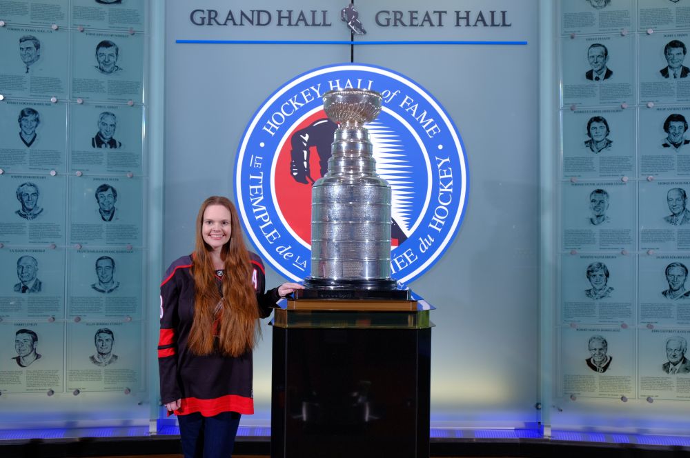 The Great Hall of the HHOF. The Stanley Cup stands tall on a base. A short woman with long hair has her hand on the pedestal.