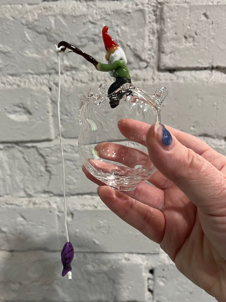 A handmade glass Christmas ornament featuring a gnome holding a fishing rod sitting on a clear glass orb. There is a purple fish bead hanging from a string tied to the rod. Bonus glimpse at my woefully chipped nail polish. 