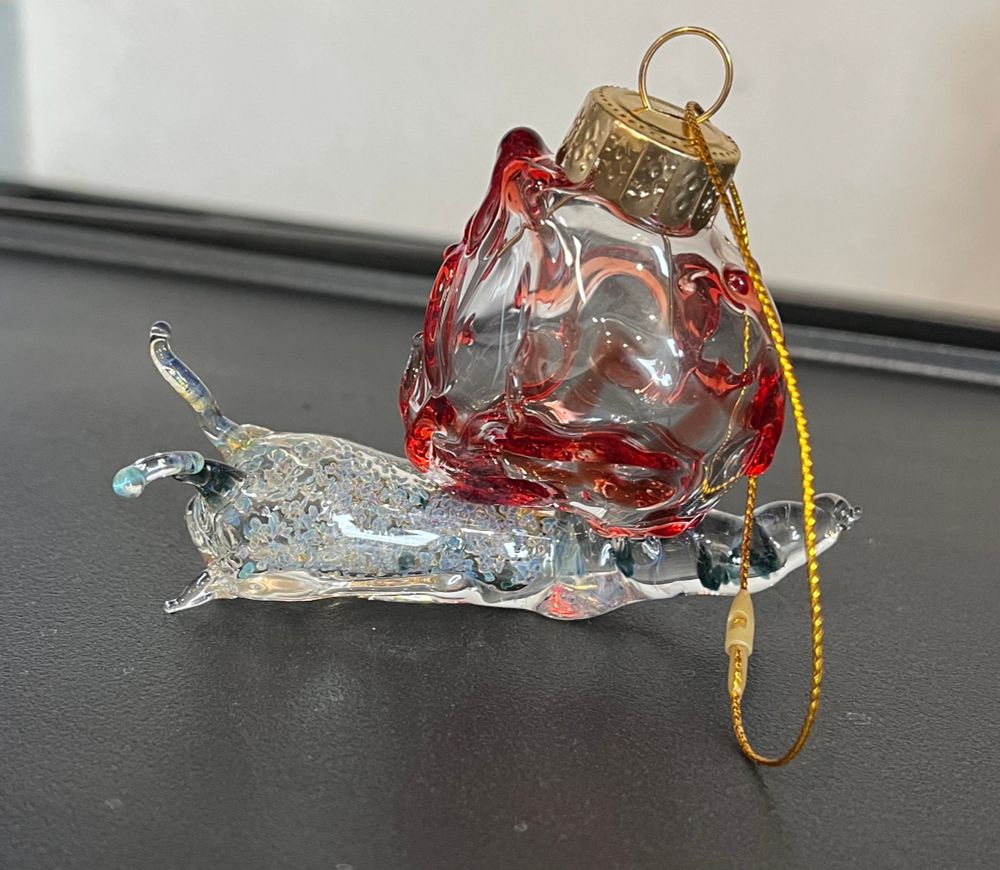 Side view of a glass snail Christmas ornament with a speckled body and a red swirly shell. It is topped by a gold ornament cap and gold string. 