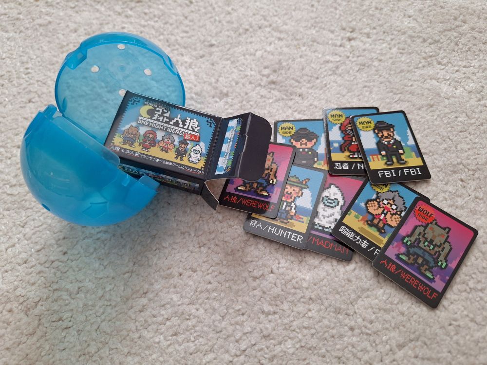 A tiny one night Werewolf game and cards spills out of an open gachapon capsule
