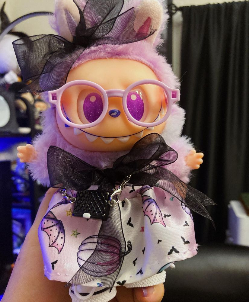 A  purple and pink stuffed monster doll dressed in a lavender Halloween dress and wearing lavender plastic glasses.